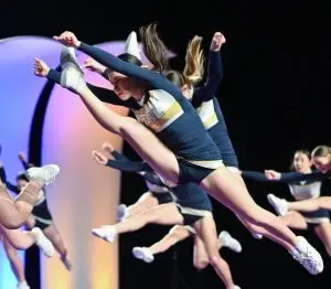 Une jeune cheerleader appartenant à Cheer Québec en uniforme bleu marine et doré exécute un saut acrobatique sur scène, avec une concentration visible sur son visage. En arrière-plan, d'autres cheerleaders participent à la performance, ajoutant de l'énergie à l'ensemble de la routine. La scène est illuminée par des lumières colorées, créant une ambiance dynamique.