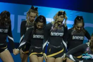 À propos cheerleading québec : Un groupe de cheerleaders en uniforme noir et doré portant le nom "Intrépides" se tient la main et regarde vers le bas sur scène. Leur expression est sérieuse et concentrée, tandis qu'elles se préparent pour leur performance. Leurs costumes scintillants ajoutent une touche de glamour à l'ambiance.