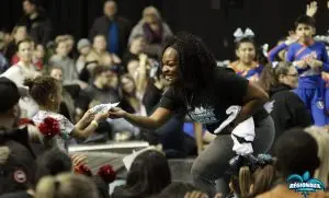 Entraîneuse de cheerleading (cheer québec) interagissant avec une jeune athlète lors d’un événement, entourée de spectateurs et d'autres participants en arrière-plan.