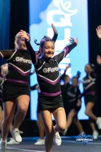 À propos le cheerleading au Québec : Une équipe de jeunes cheerleaders en uniforme noir et violet, souriants et en mouvement sur scène, lors d'une performance. L'un des cheerleaders au premier plan affiche une expression joyeuse, avec les bras levés et une coiffure dynamique. En arrière-plan, d'autres membres de l'équipe se préparent à participer à la routine.